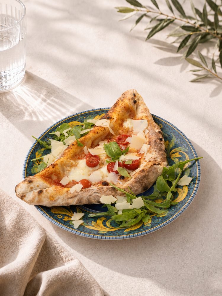 Halbmond aus Pizzateig: eine Hälfte gefüllt, die andere belegt mit Tomatensoße, Mozzarella, Cherrytomaten und Parmesanflocken, auf Rucola serviert